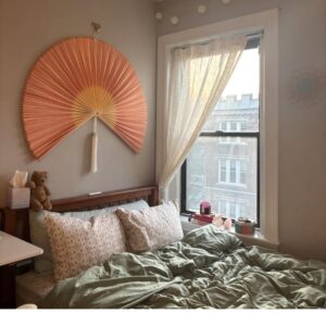 Pink bamboo wall fan decor hanging above a bed near a window adding a soft and natural accent to a modern bedroom
